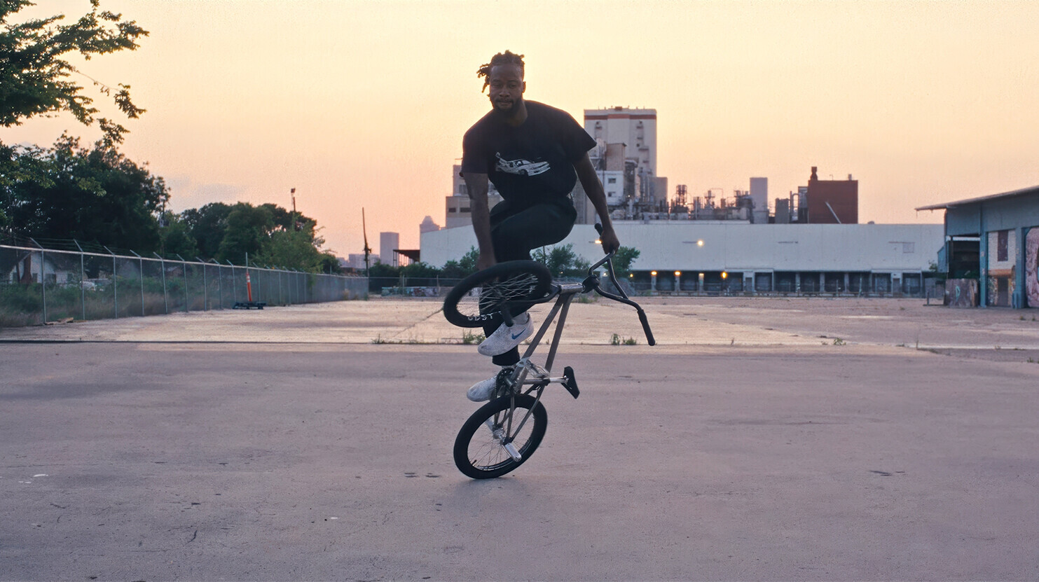 Omari Cato f&uuml;hrt bei Sonnenuntergang in einer st&auml;dtischen Industriegegend einen Flatland-BMX-Trick vor.