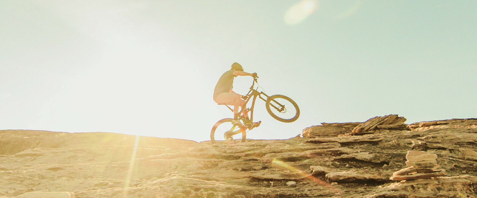 Mountainbiker fährt einen felsigen Anstieg hinauf bei Sonnenuntergang, mit sichtbaren Sonnenstrahlen im Hintergrund.