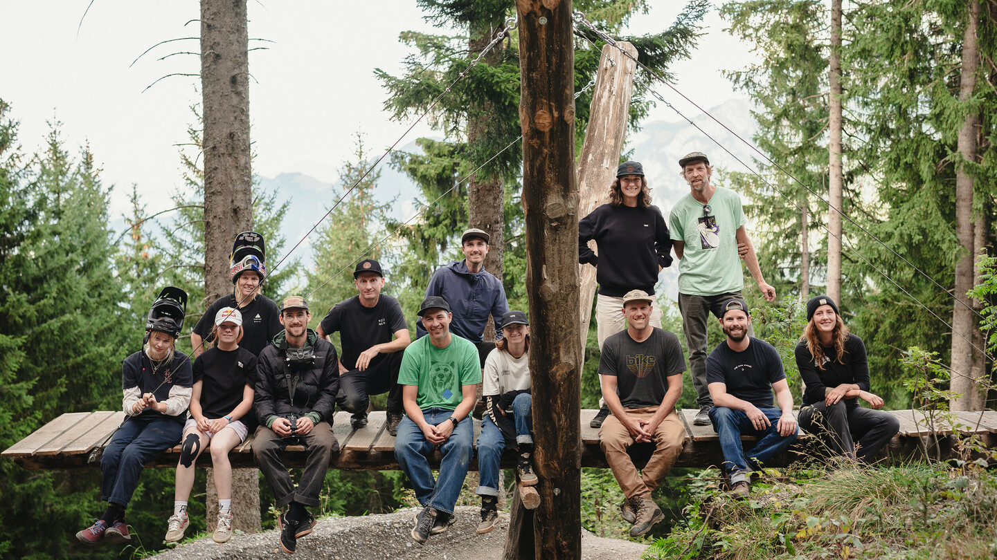 Eine Gruppe von Mountainbikern und Crewmitgliedern posiert gemeinsam auf einer Holzplattform im Wald. Die Personen tragen sportliche Kleidung und Helme, und lächeln in die Kamera.