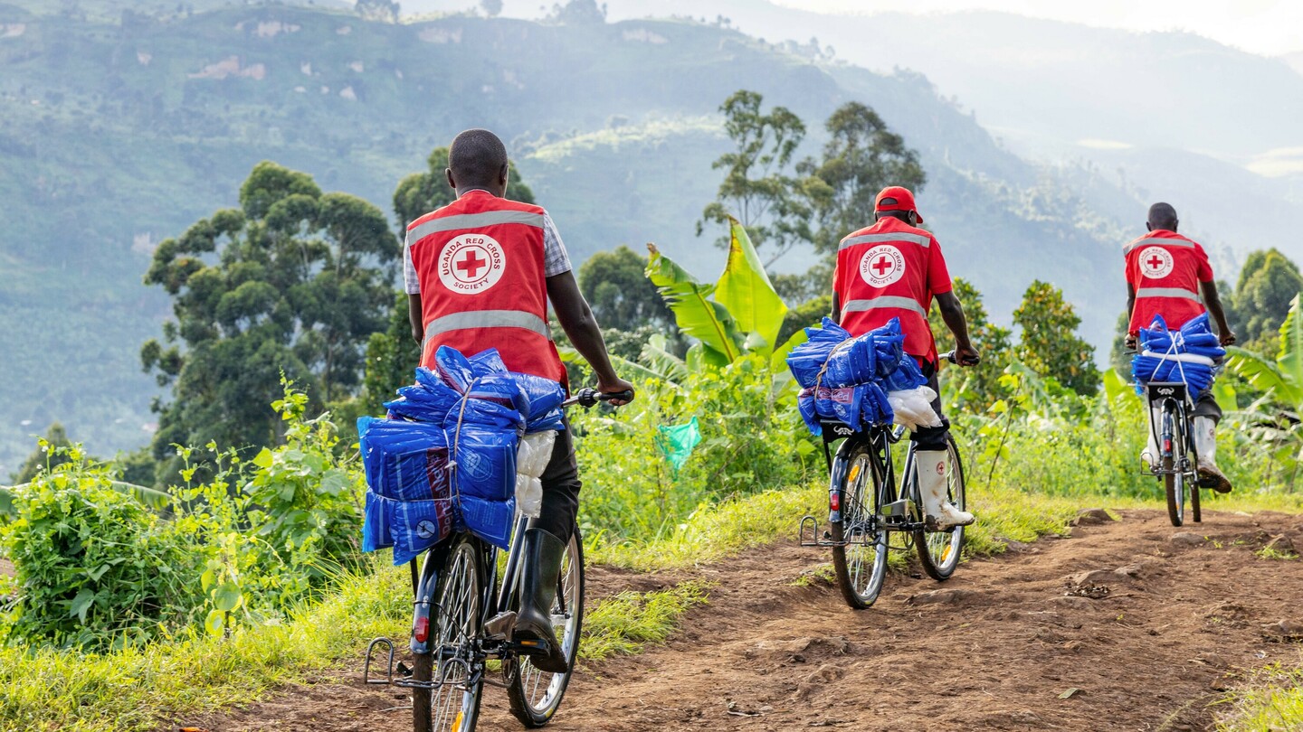 Drei Männer in roten Westen mit dem Logo des Uganda Red Cross Society fahren auf Fahrrädern beladen mit blauen Paketen auf einem ländlichen Weg durch grüne Landschaft.