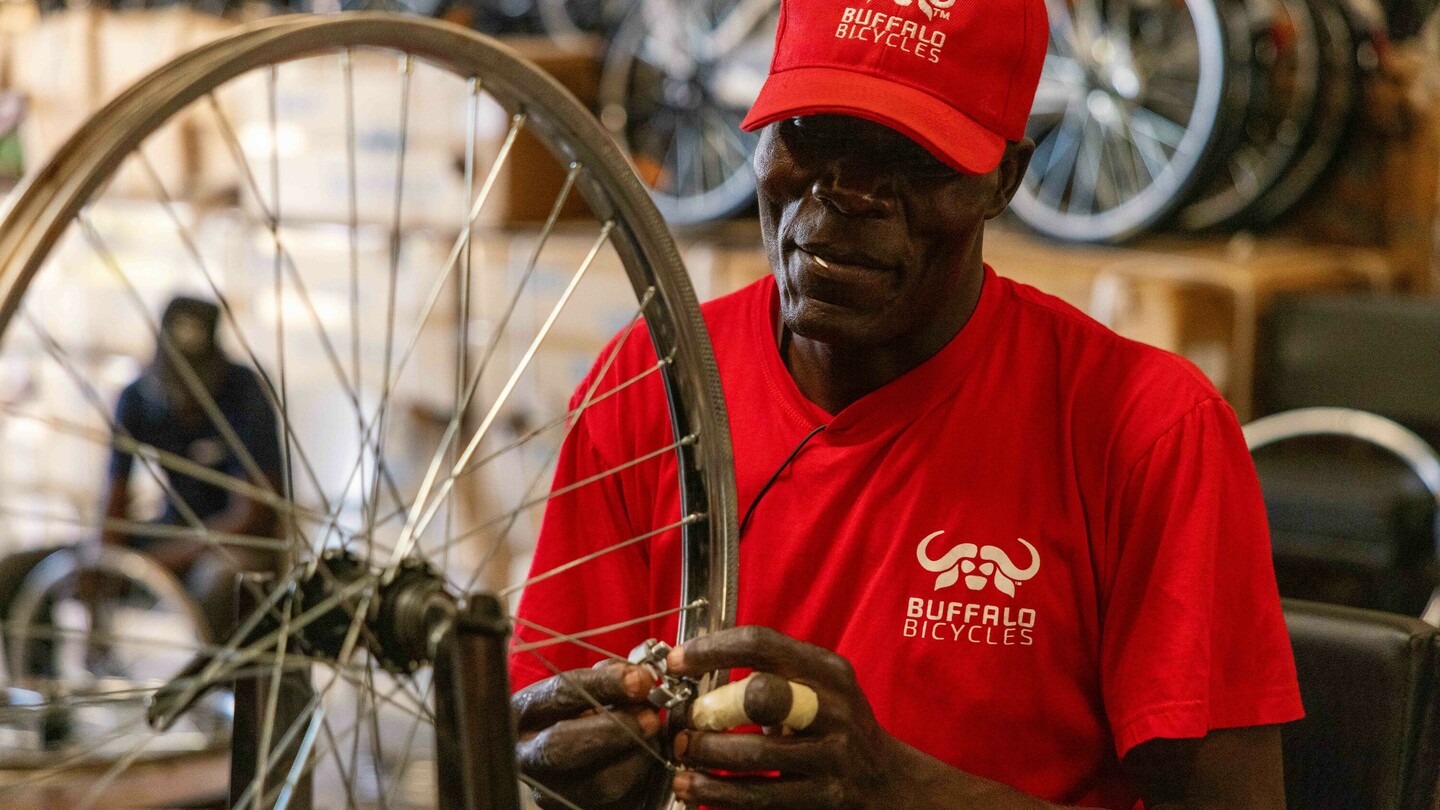 Ein Mann in rotem Shirt und Kappe von Buffalo Bicycles arbeitet an einem Fahrradreifen in einer Werkstatt. Im Hintergrund sind verschwommen gestapelte Fahrräder und Kartons zu sehen.