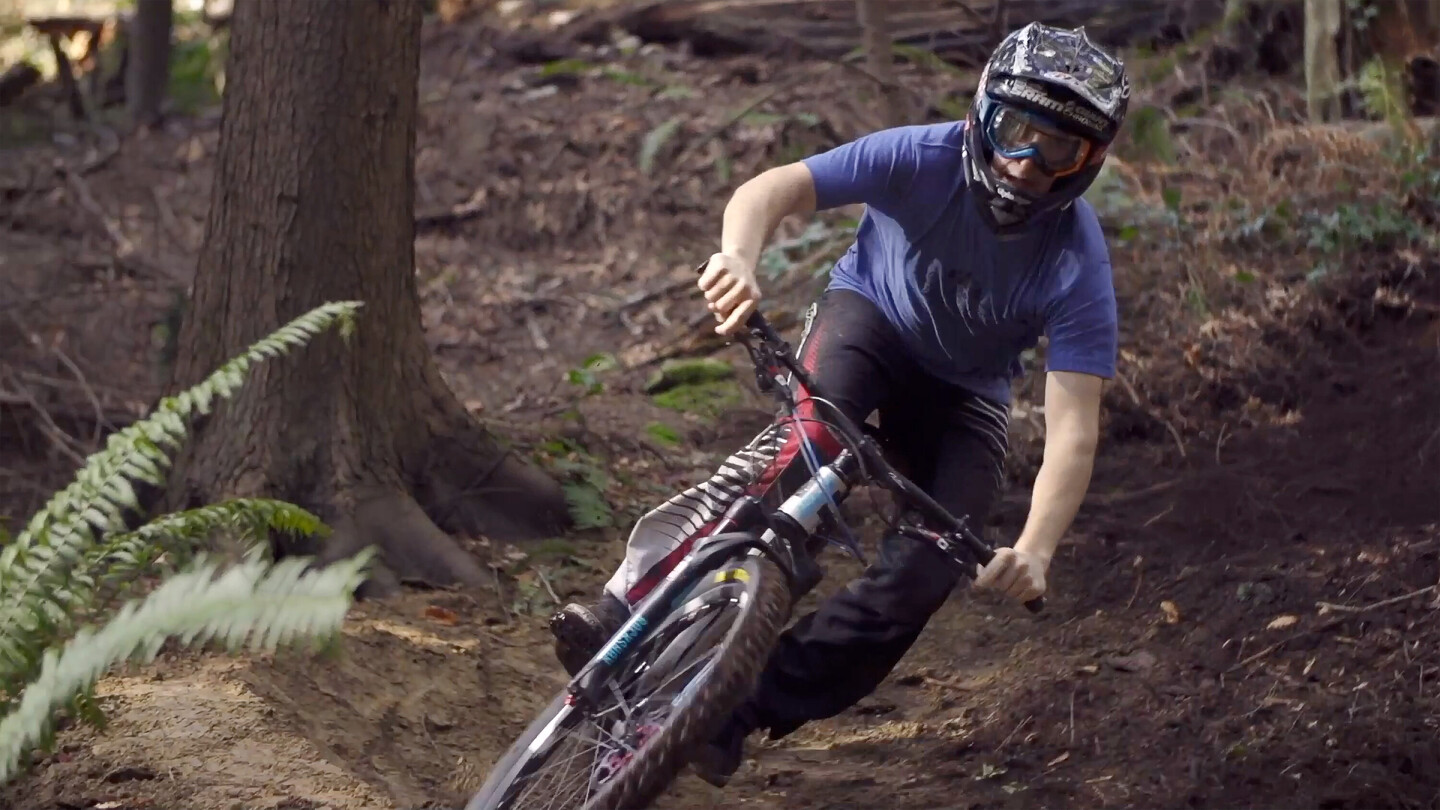 Ein Mountainbiker mit Helm und Brille fährt in einer Kurve durch einen Wald. Der Fahrer trägt ein blaues T-Shirt und schwarze Hosen und steuert das Fahrrad präzise durch den unebenen, erdigen Untergrund.