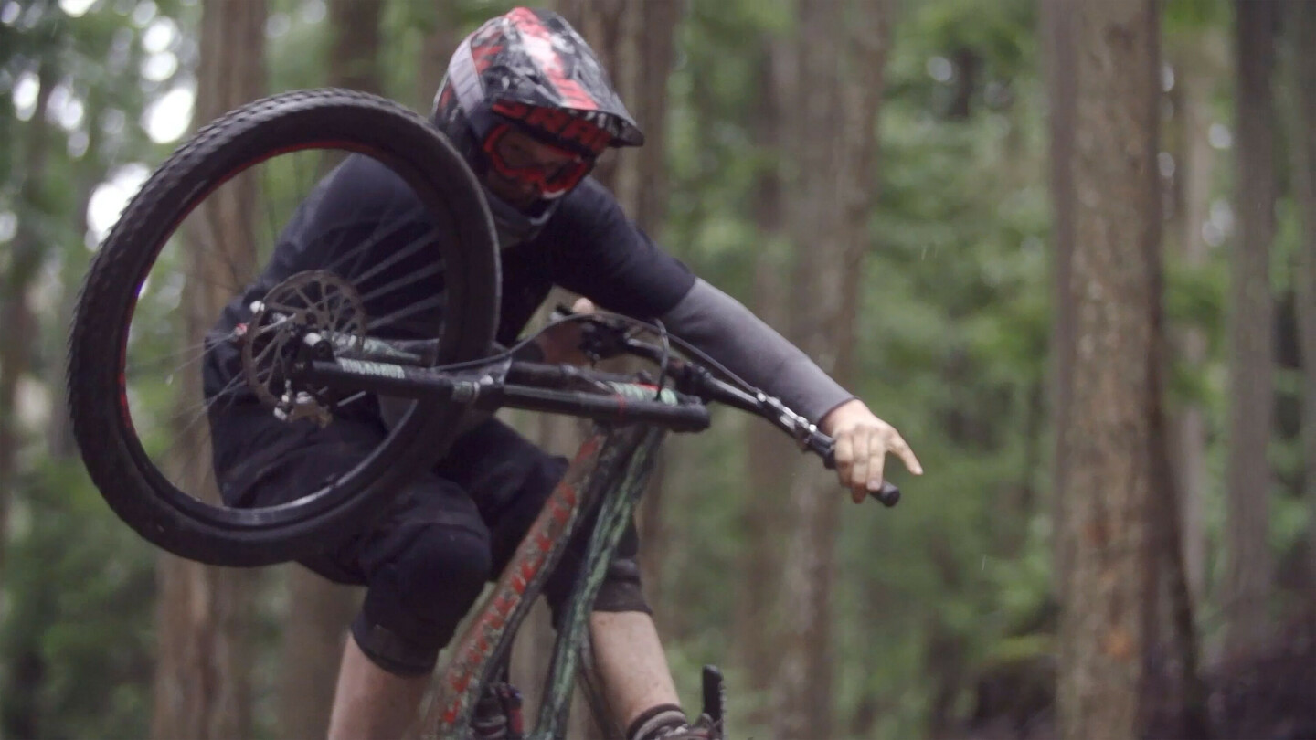 Mountainbiker mit Helm und Schutzbrille macht einen Sprung oder Trick in einem Wald, mit angehobenem Vorderrad.