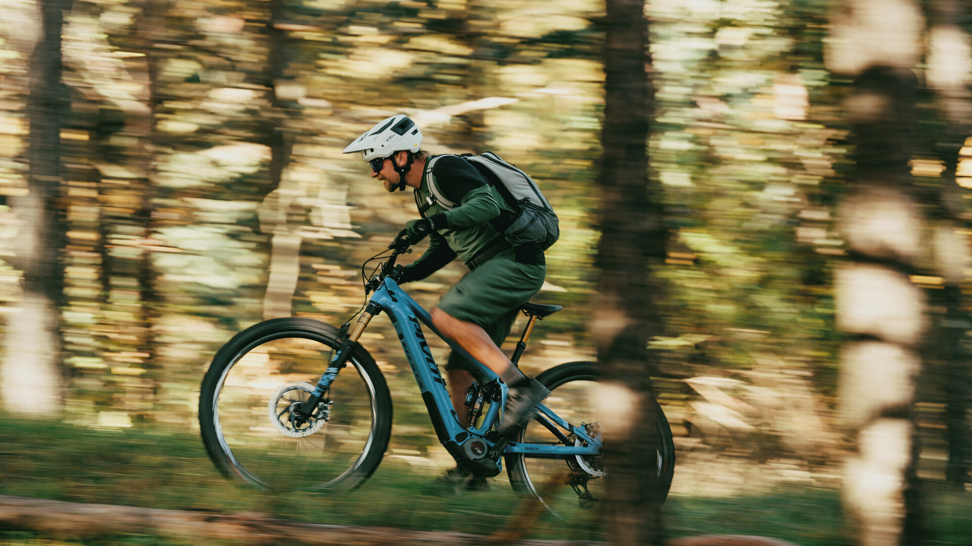 Mountainbiker mit Helm und Rucksack fährt mit hoher Geschwindigkeit auf einem blauen Fahrrad durch einen Wald.